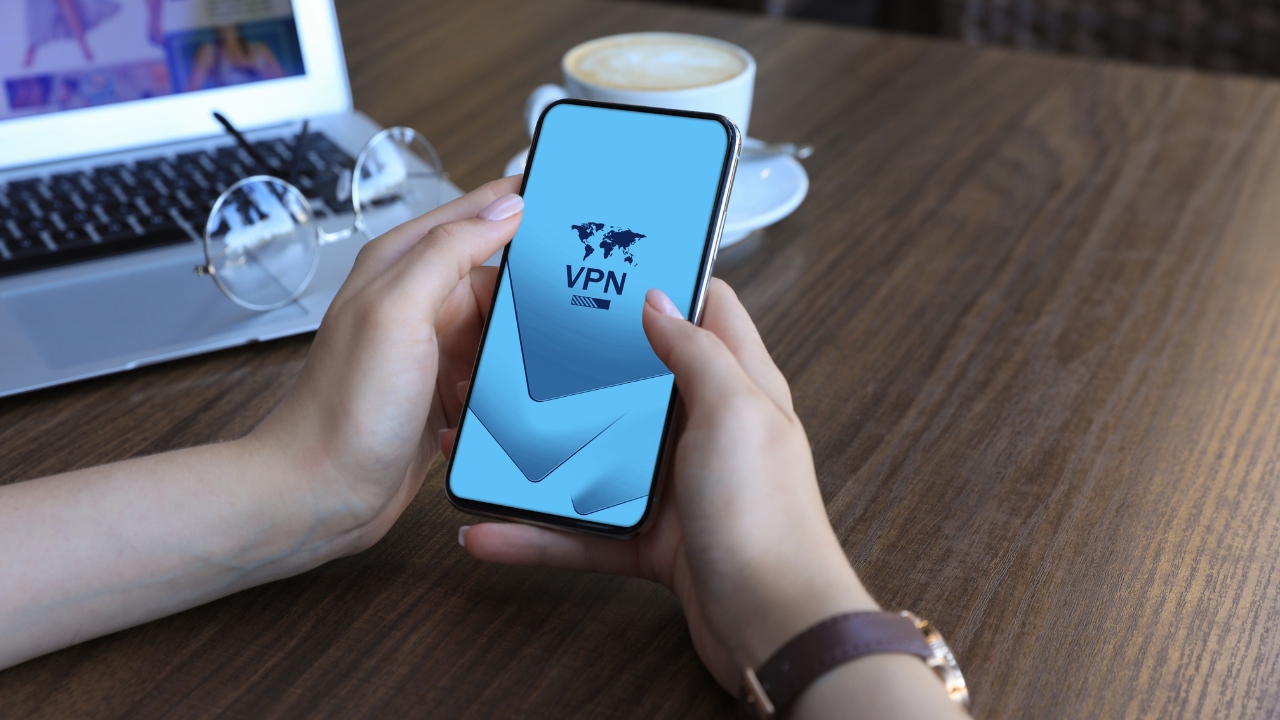 Person holding a smartphone with a VPN app open on the screen near a laptop and coffee cup
